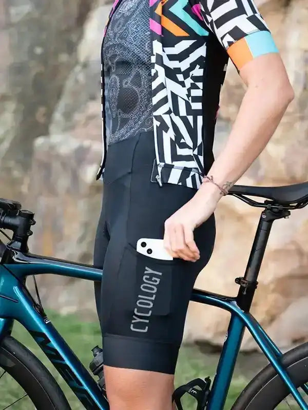 Cyclist in geometric print jersey and black cycling shorts with phone pocket, standing beside teal road bike