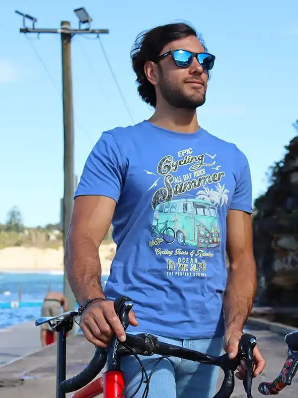 Man wearing blue cycling t-shirt with van graphic, holding bike by beach, summer ride apparel