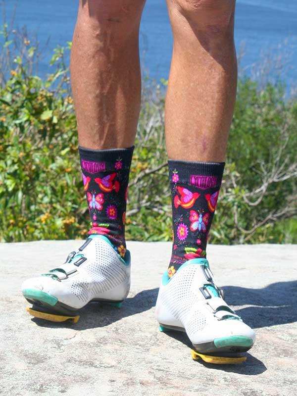 Colorful floral cycling socks with butterflies, paired with white road bike shoes, outdoors near water