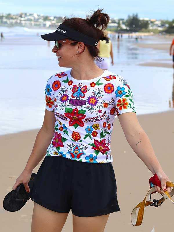 Woman in colorful floral cycling jersey and black shorts at beach, holding sandals and visor