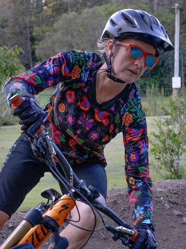 Woman cycling in colorful floral long-sleeve jersey, helmet, and sunglasses on dirt trail