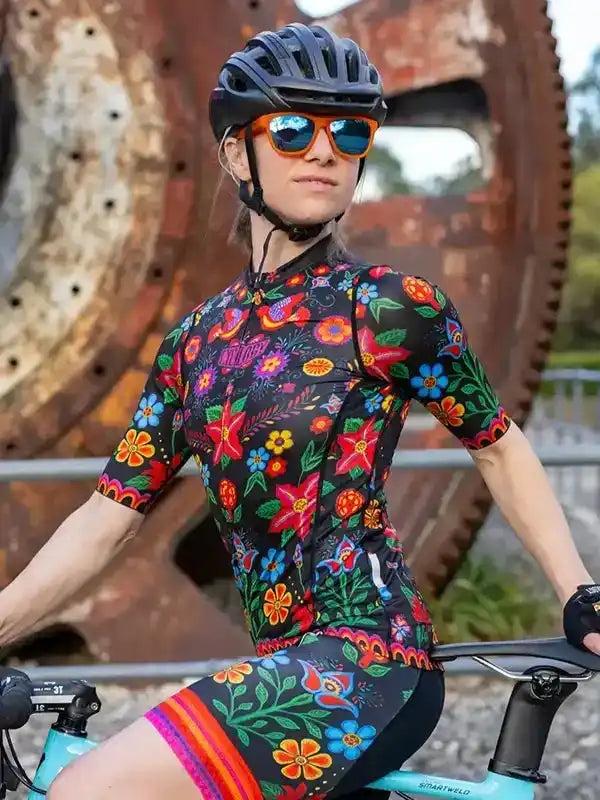 Woman in colorful floral cycling jersey and shorts, wearing helmet and sunglasses, on bike near rustic gear sculpture
