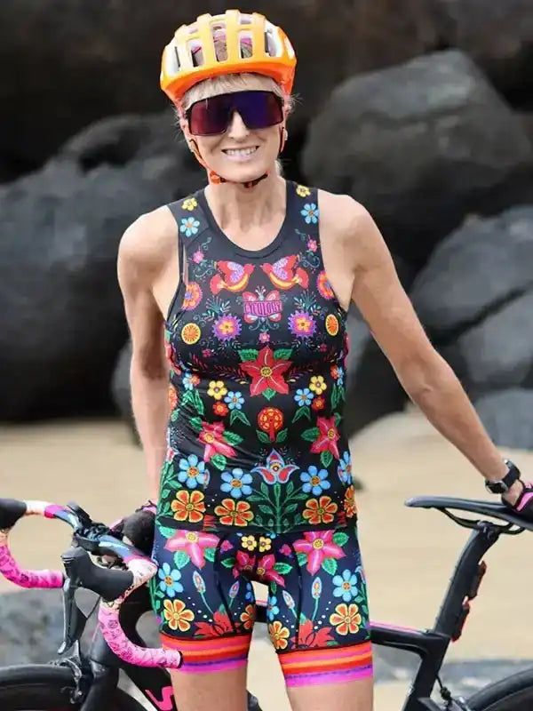 Woman in floral cycling jersey and shorts with orange helmet, standing by bike on rocky beach