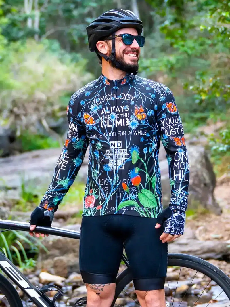 Man in floral cycling jersey and helmet, standing with road bike in forest setting, wearing sunglasses and gloves