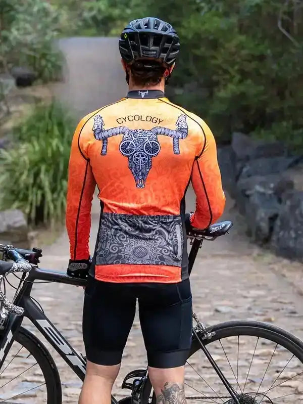Man wearing orange long-sleeve cycling jersey with bull skull design, standing with road bike on trail