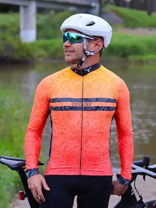 Man wearing orange patterned cycling jersey with 'CYCOLOGY' logo, helmet, and sunglasses near water