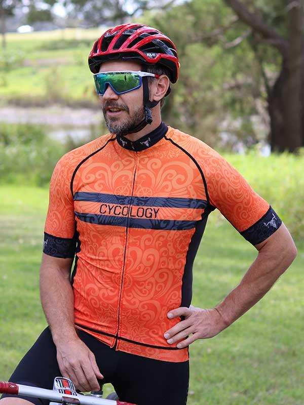 Man wearing orange patterned cycling jersey with 'CYCOLOGY' logo, helmet, and sunglasses outdoors