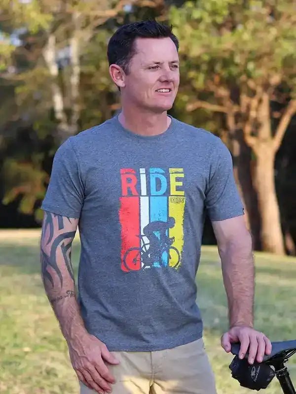 Man wearing gray cycling t-shirt with colorful 'RIDE' graphic and cyclist design, outdoors near bike