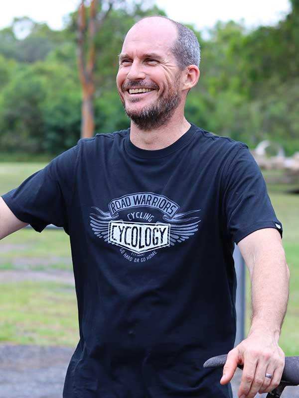 Man wearing black 'Road Warriors Cycling' t-shirt outdoors, smiling near bike