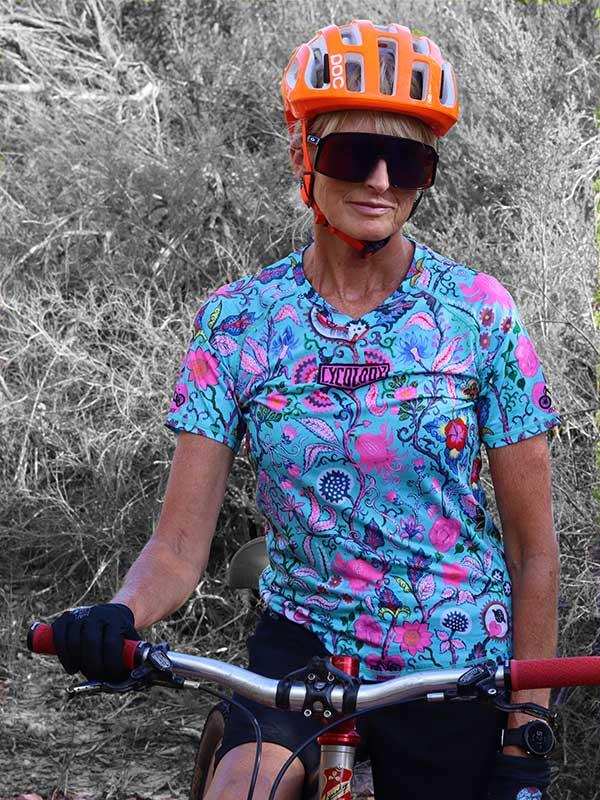 Woman cycling in colorful floral jersey, orange helmet, and sunglasses on trail