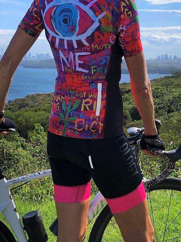 Colorful graphic cycling jersey with eye design, black shorts, pink leg bands, scenic waterfront backdrop