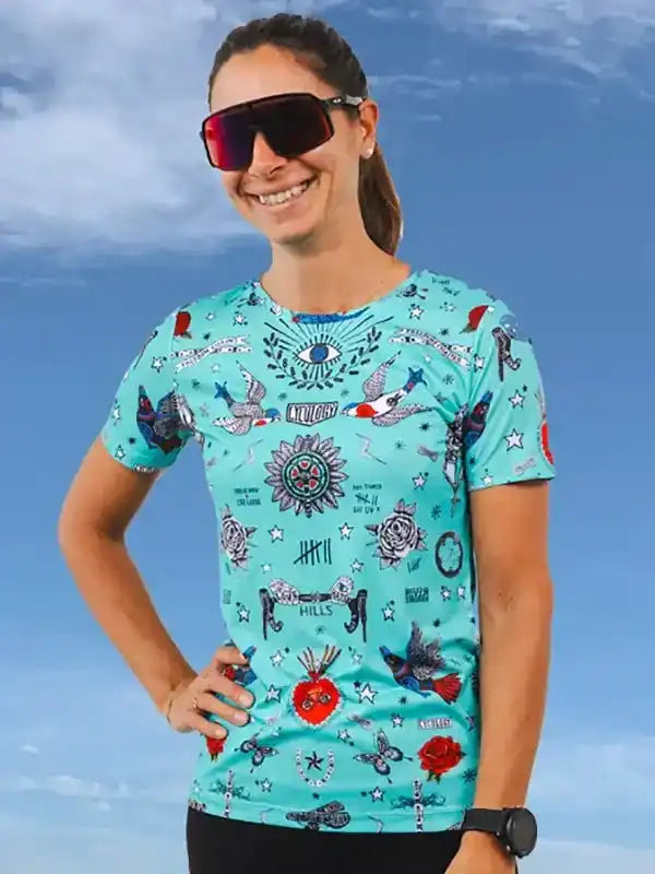 Woman wearing vibrant turquoise cycling jersey with tattoo-style graphics, sunglasses, and sporty pose against sky