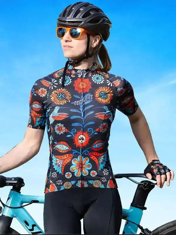 Woman in colorful floral cycling jersey, helmet, and sunglasses, holding bike against blue sky