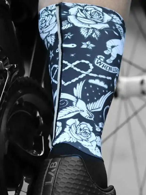 Cycling leg warmer with white rose and swallow tattoo design on dark blue fabric, worn with cycling shoe