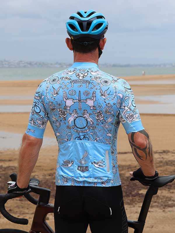 Man in light blue patterned cycling jersey and helmet, standing with bike on beach