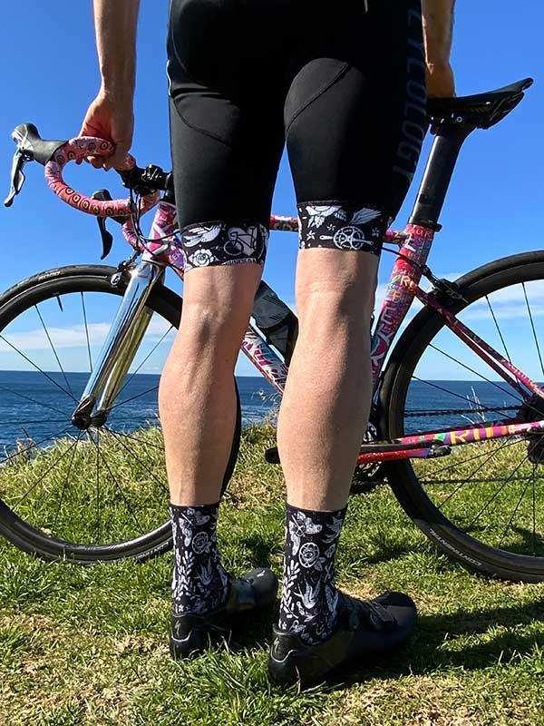 Cyclist wearing black cycling shorts with floral patterned cuffs and matching socks, standing beside a colorful road bike by the ocean.