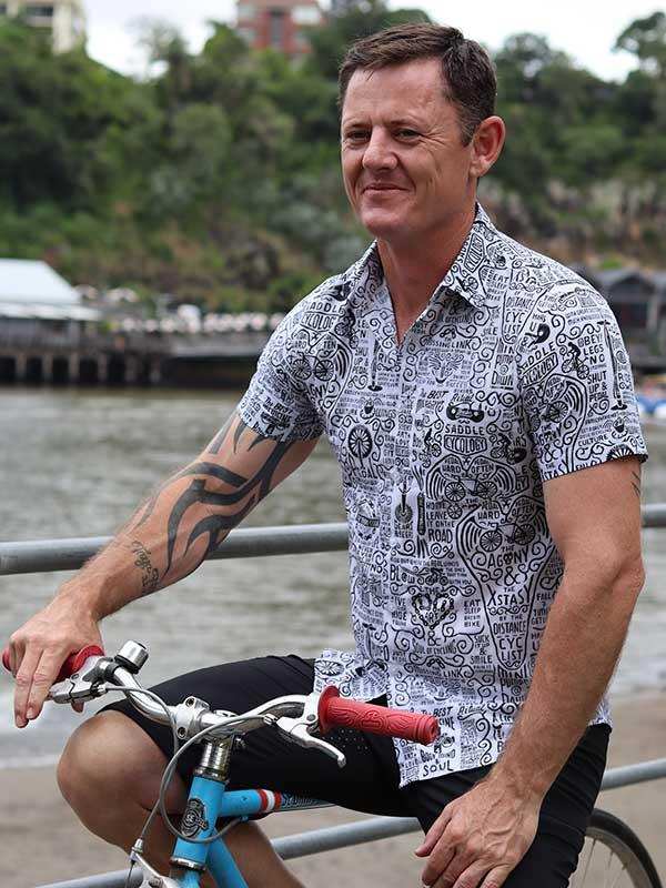 Man wearing graphic print cycling shirt and black shorts on blue bike by water